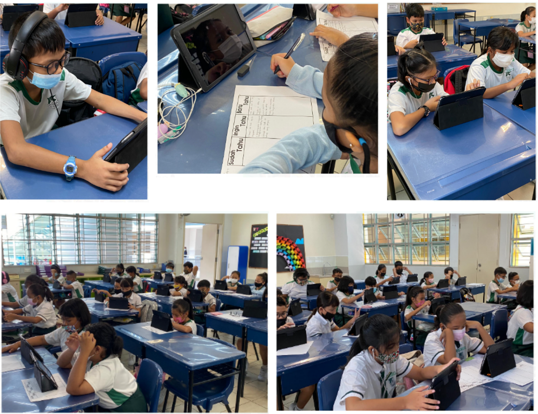 Students wearing masks sit at blue desks in a classroom, using tablets and writing on worksheets.
